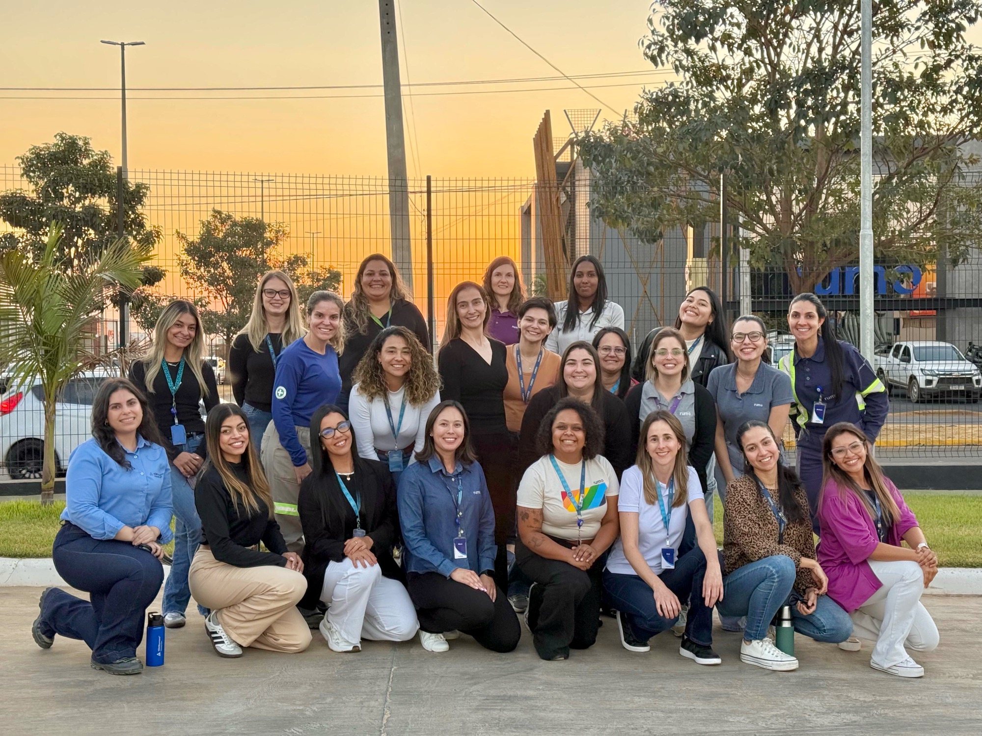Grupo de mulheres posando ao ar livre ao entardecer em frente a cerca e prédio.