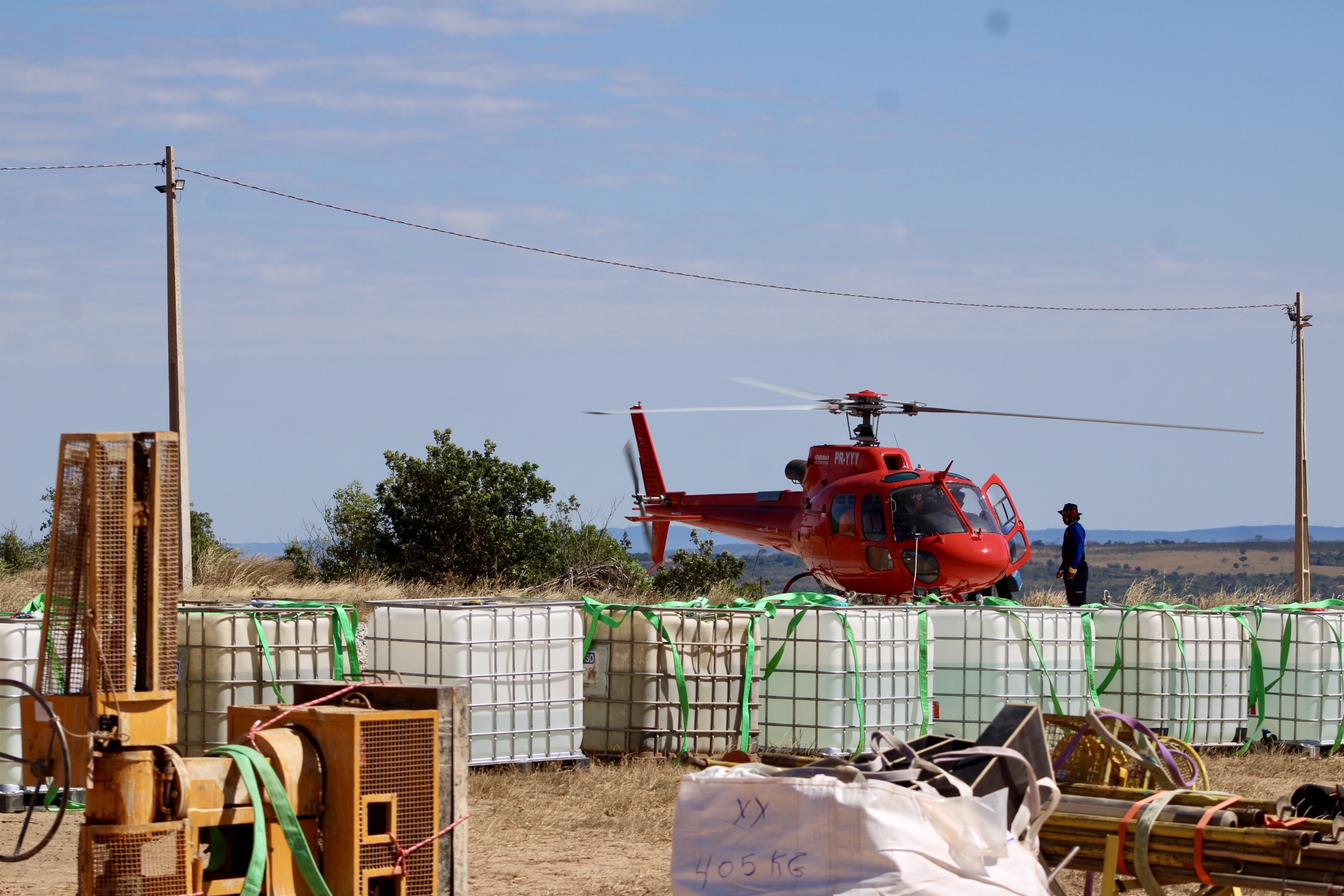 Helicóptero vermelho estacionado atrás de grandes contêineres plásticos brancos amarrados com cintas verdes e equipamentos no primeiro plano.