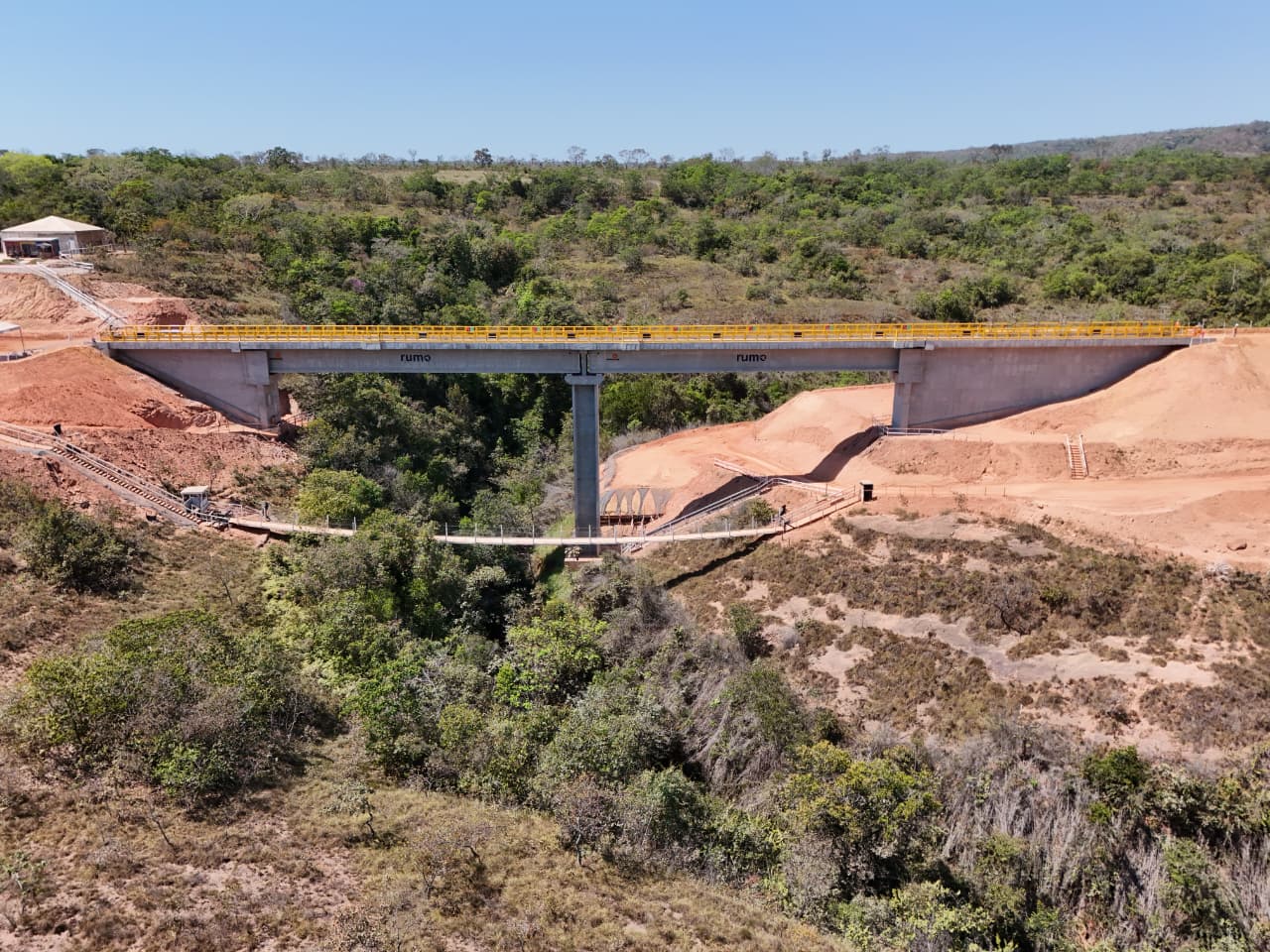 Vista aérea de ponte de concreto com guarda-corpos amarelos atravessando um vale com vegetação e áreas de terraplenagem.
