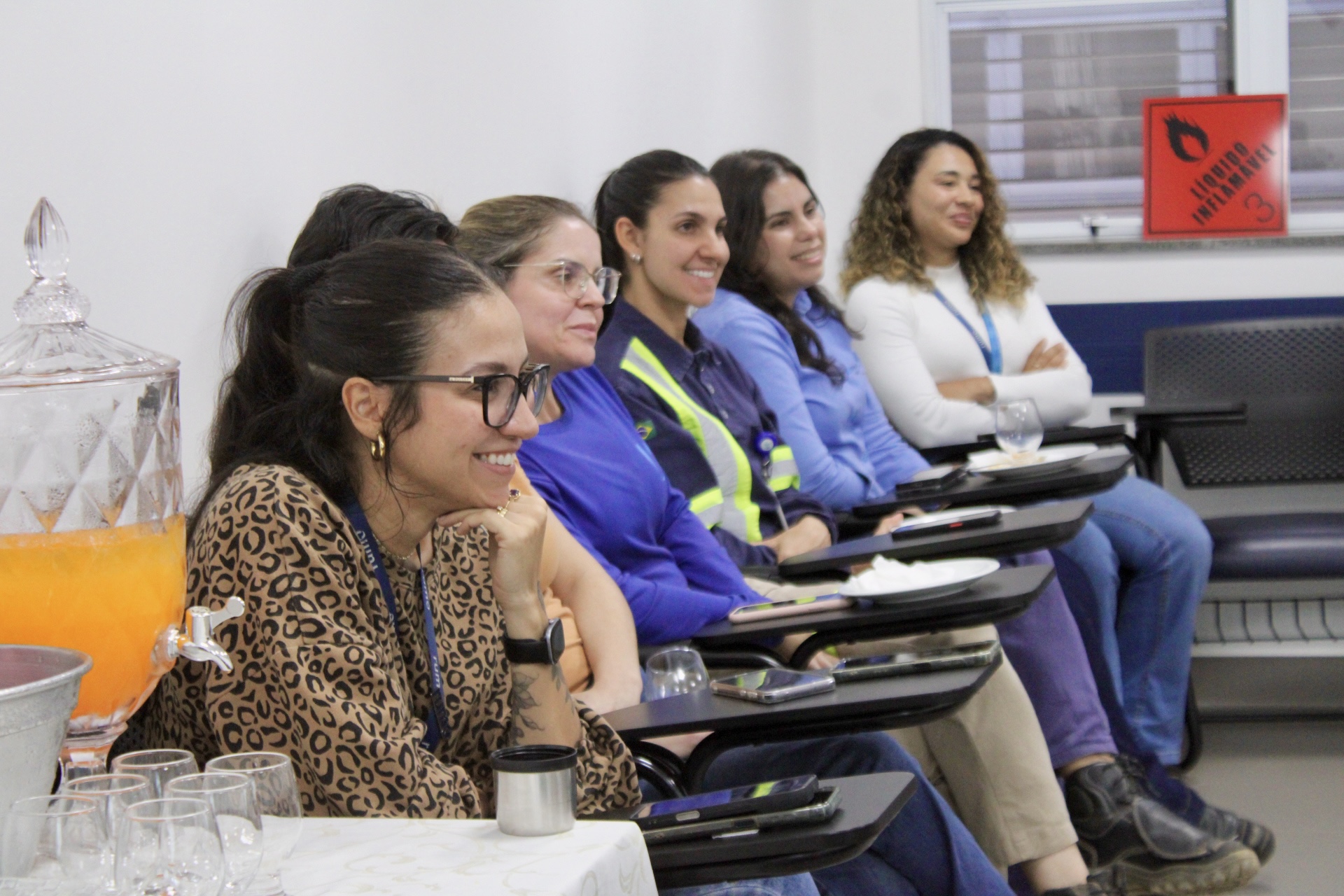 Mulheres sentadas em fila em reunião, sorrindo; jarro de suco e copos à esquerda.