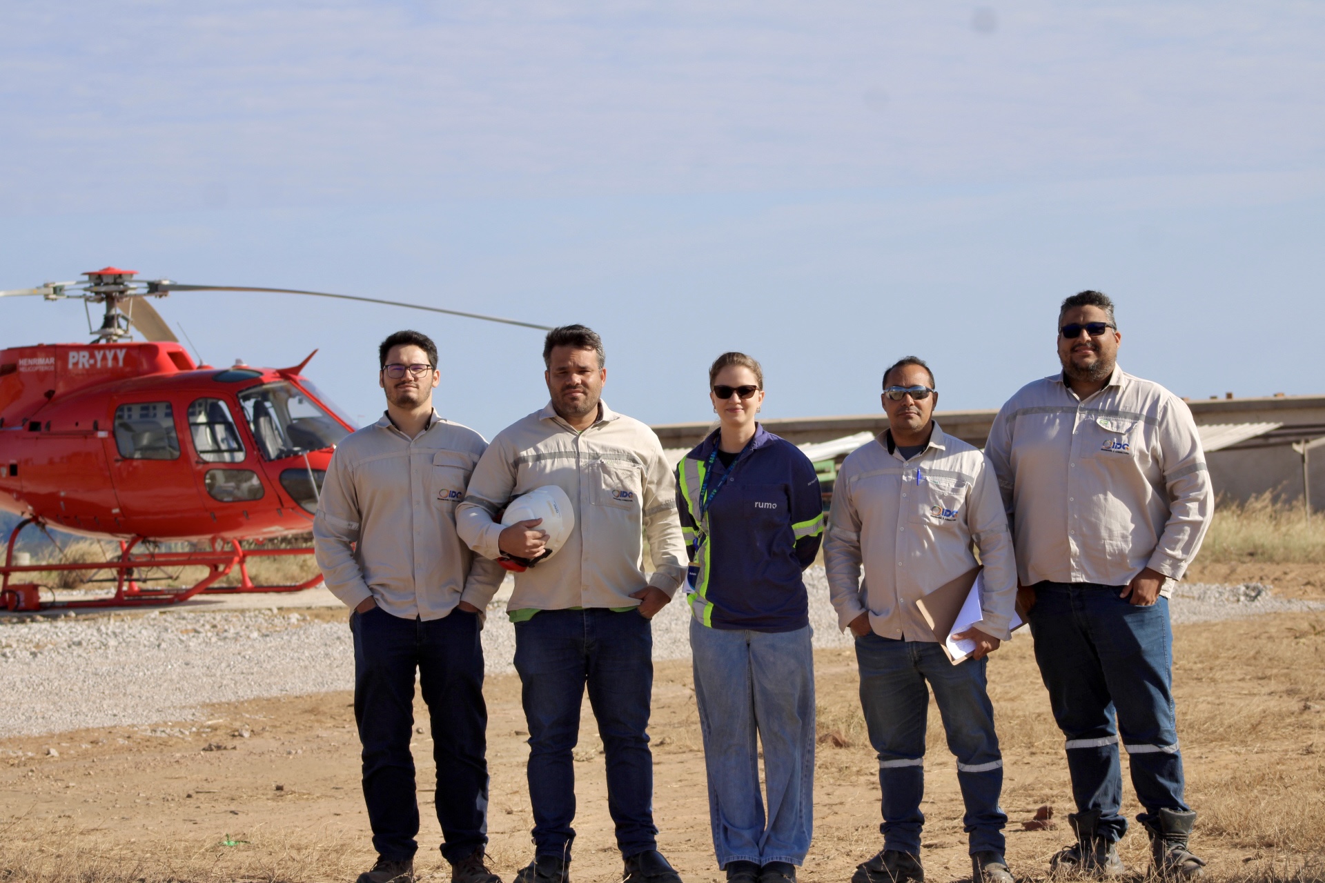 Cinco trabalhadores em pé num terreno seco, alguns com capacetes, posando à frente de um helicóptero vermelho estacionado.
