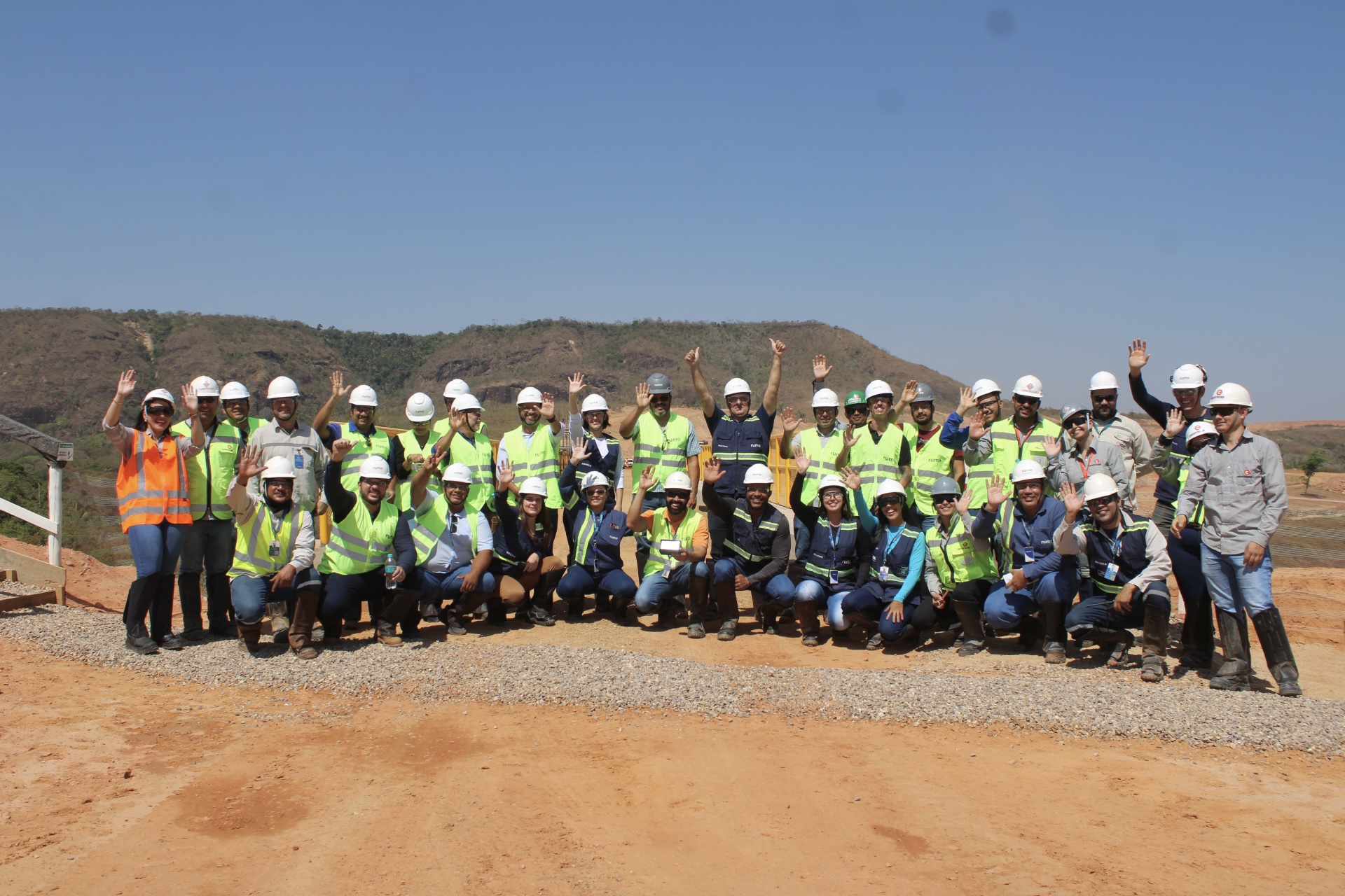 Grande grupo de trabalhadores em obra com coletes e capacetes, posando e acenando ao ar livre.