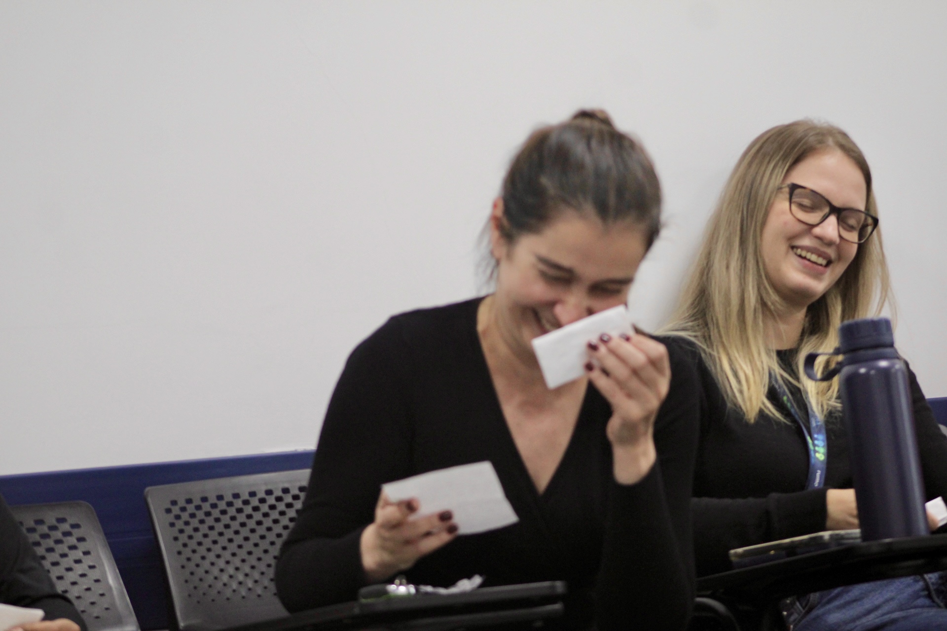 Duas mulheres sentadas sorrindo; uma limpa os olhos com um lenço enquanto segura cartões.
