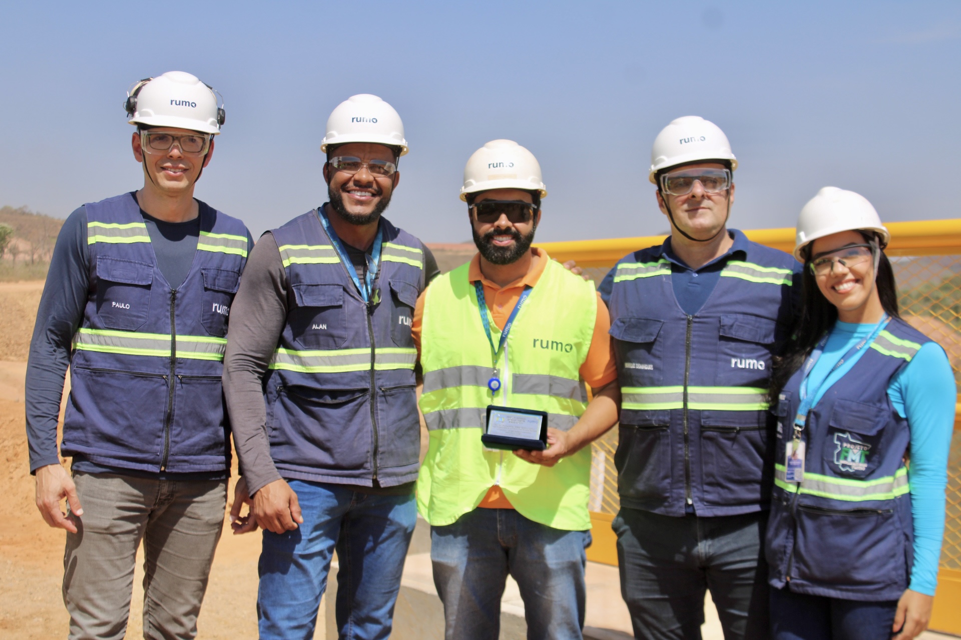 Cinco trabalhadores com capacetes e coletes posam; o do centro segura uma placa comemorativa.
