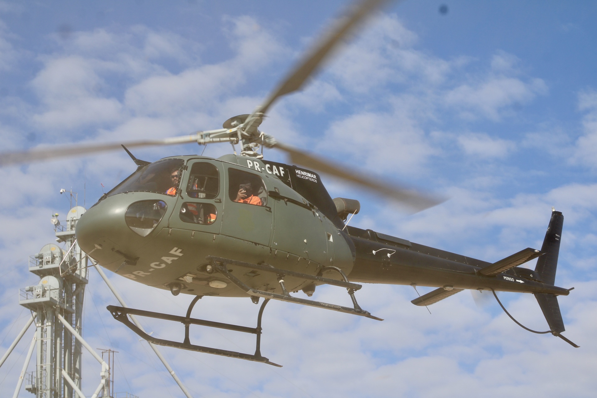 Helicóptero cinza em voo baixo contra céu azul; tripulantes com coletes laranja visíveis pela cabine.