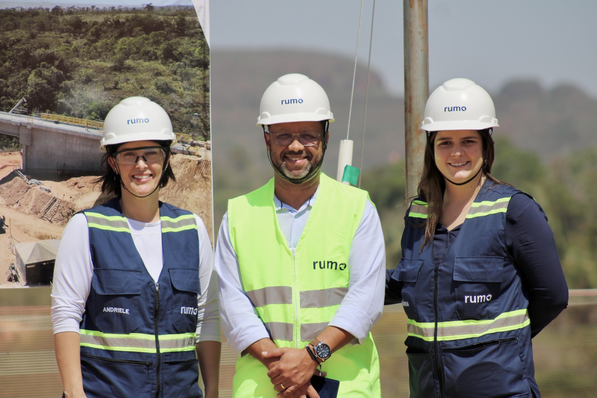 Três profissionais com capacetes e coletes posam em obra; colete central é neon com logo "Rumo".