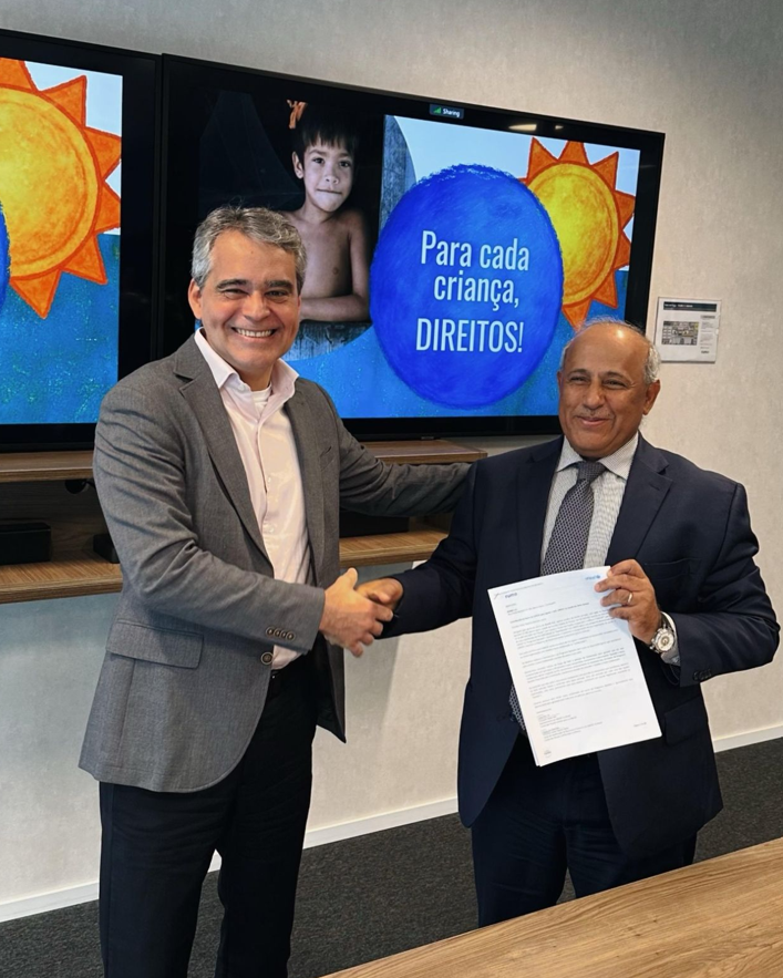Dois homens de terno apertam as mãos; um segura documento; tela ao fundo diz "Para cada criança, DIREITOS!".